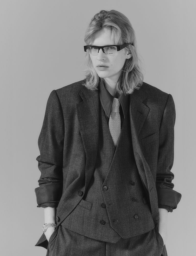 Monochrome portrait of a young adult in a suit, vest, dress shirt, tie, and glasses, with a neutral expression against a gray background.