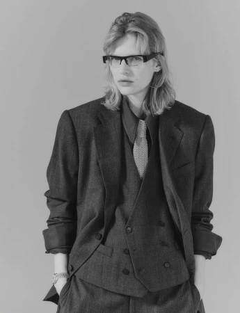 Monochrome portrait of a young adult in a suit, vest, dress shirt, tie, and glasses, with a neutral expression against a gray background.