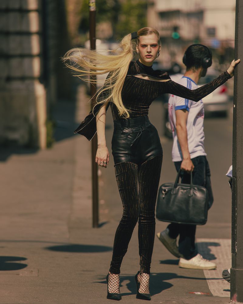 Fashionable woman in black outfit poses confidently on a city street, with a blurred pedestrian carrying a black bag in the background.