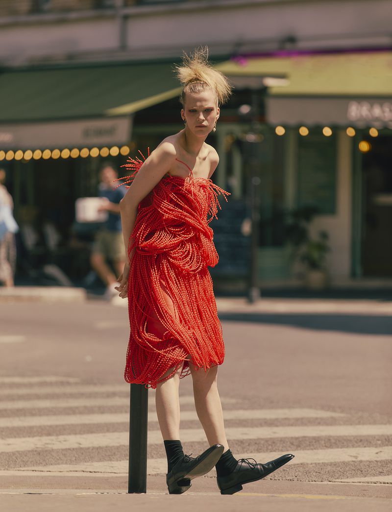 An adult female model in a red dress stands on a crosswalk in an urban area, with buildings and trees in the background on a sunny day.