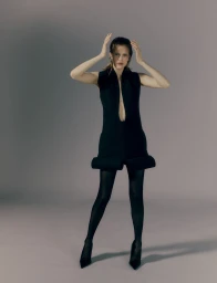 Model in a black sleeveless dress and dark tights posing against a plain grey studio backdrop.