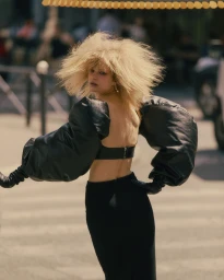 Blonde model with voluminous hair wearing dramatic black puff sleeves and a backless top posing on a sunlit street.