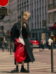 Model in a black coat and red skirt standing on a city street for Special Mode by Elle France