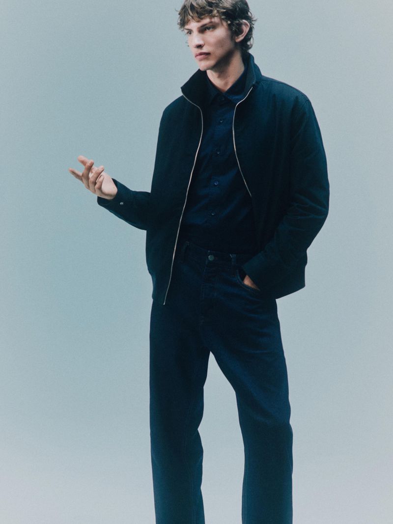 Studio shot of a young adult male model in a navy jacket and pants against a light blue backdrop, showcasing fashion and style.