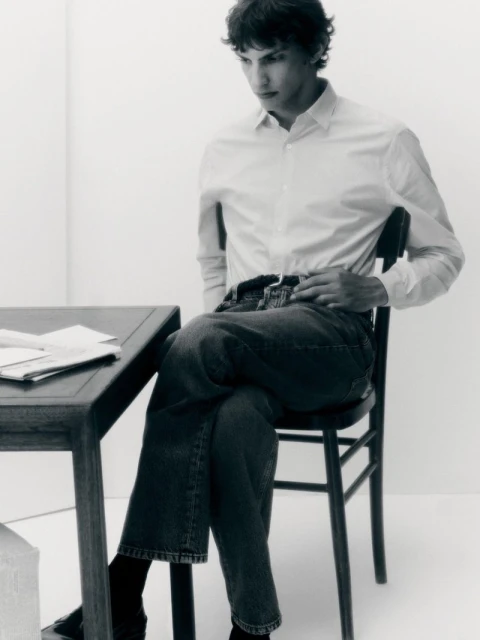 Adult male sits at a dark wood desk with papers in a minimally decorated room. He wears jeans and a button-down shirt.