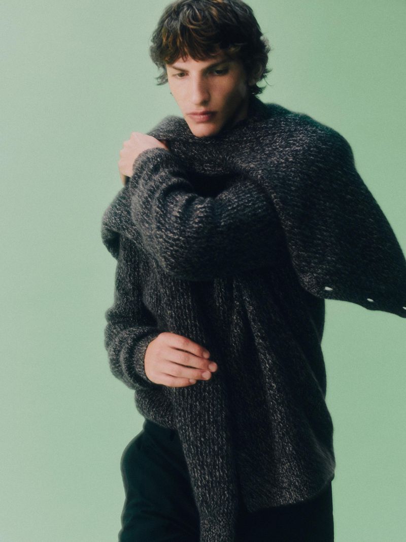 Young man wearing a textured charcoal grey knit sweater draped with a matching jumper against a pale green studio background.