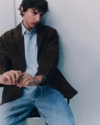 Man wearing a brown blazer, light blue shirt, and blue jeans against a neutral studio background.