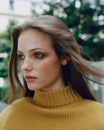 Portrait of a young woman with long hair in a yellow turtleneck sweater against a blurred background of trees and a building.