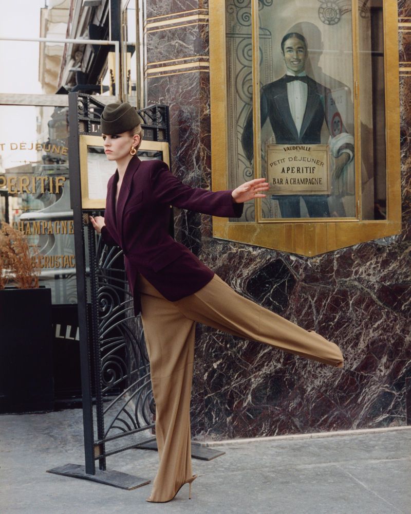 Fashion model in a burgundy blazer, tan slacks, and green hat posing in front of a marble building with a vintage advertisement.