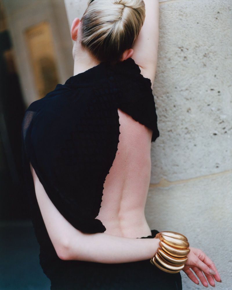 A woman with blonde hair is shown from the back, wearing a black top, back to the viewer, with a gold cuff bracelet on her wrist.