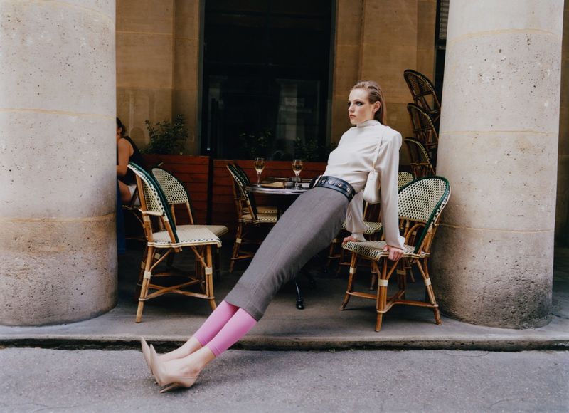Fashion photography of a woman in a white blouse and gray skirt leaning between pillars and chairs on a patio.