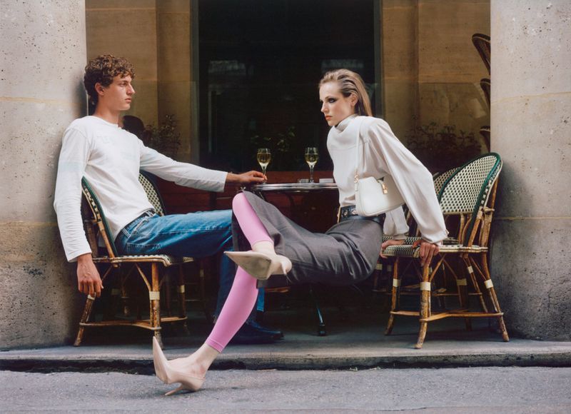 A couple poses in front of an outdoor cafe. The man wears blue jeans and the woman a white shirt, gray trousers, and nude heels.