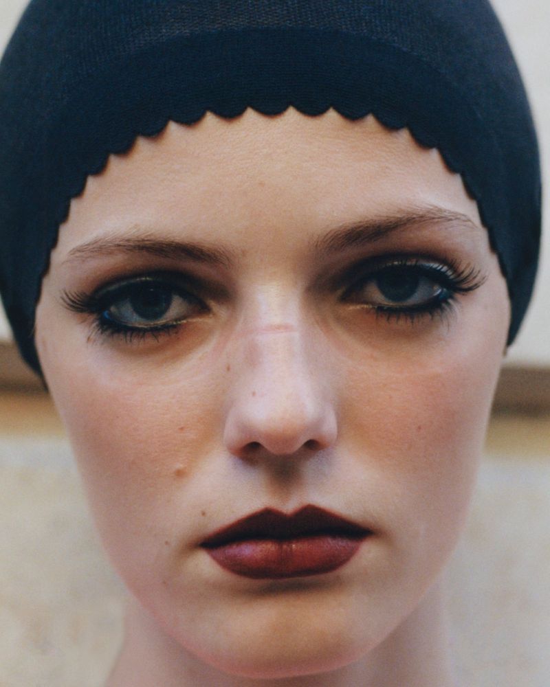 Close-up portrait of a young adult female with black cap, dark lipstick, and dramatic eye makeup, gazing directly into the camera.