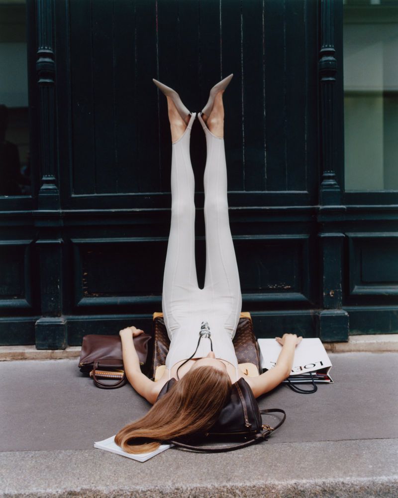 Upside-down view of a woman lying on a sidewalk with legs and high heels against a dark door; handbags and shopping bag visible.