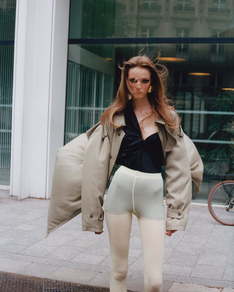 Fashionable redhead woman in black top and trench coat posing outdoors near modern building with a bicycle parked nearby.