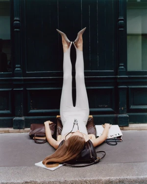 Woman in a light grey catsuit and heels lies on a sidewalk with legs raised straight up against a dark paneled wall next to handbags.