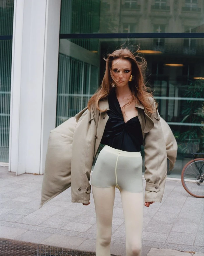 Model wearing an oversized beige trench coat, asymmetrical black top, and pale leggings on a city street for Vogue China.