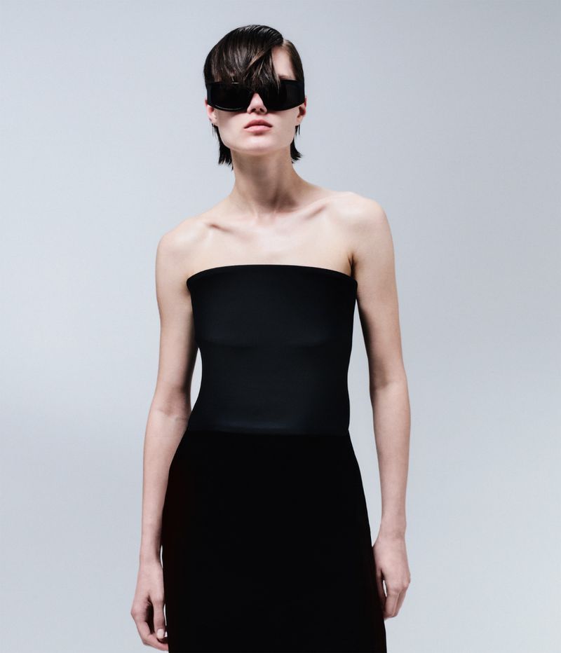 Model with dark short hair wearing oversized black shield sunglasses and a strapless black top against a grey seamless backdrop.