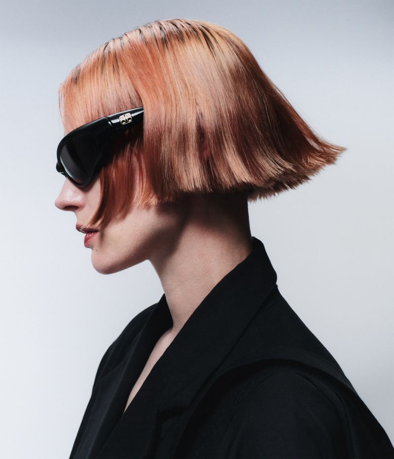 A woman with a short bob hairstyle and black sunglasses, shot in profile against a plain background, wearing a black blazer.