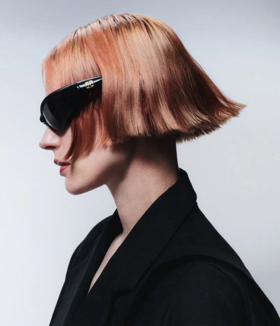 A woman with a short bob hairstyle and black sunglasses, shot in profile against a plain background, wearing a black blazer.