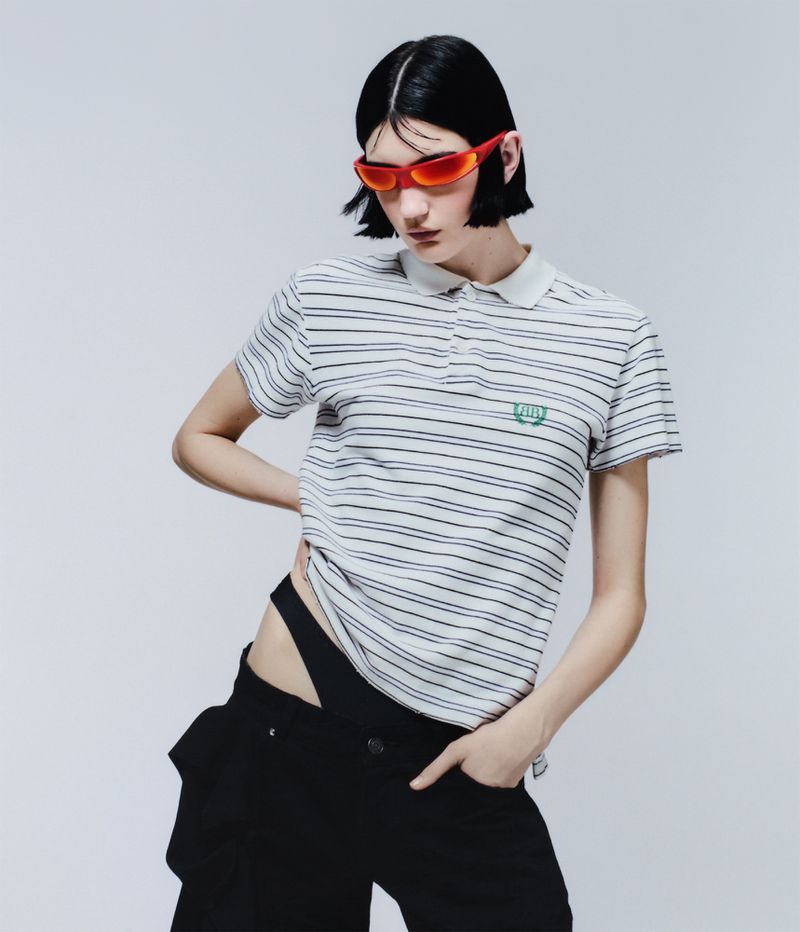 Striking fashion shot of a young woman with short black hair, red sunglasses, and a striped polo shirt against a plain white backdrop.