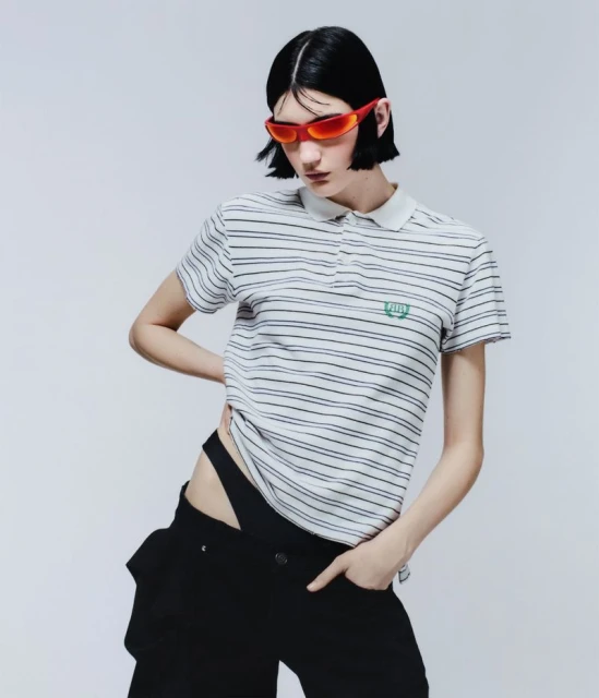 Striking fashion shot of a young woman with short black hair, red sunglasses, and a striped polo shirt against a plain white backdrop.