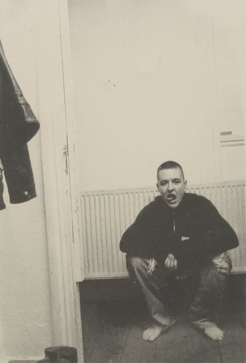 Black and white portrait of a squatting man with a shaved head sticking his tongue out in an indoor setting with a hanging jacket.