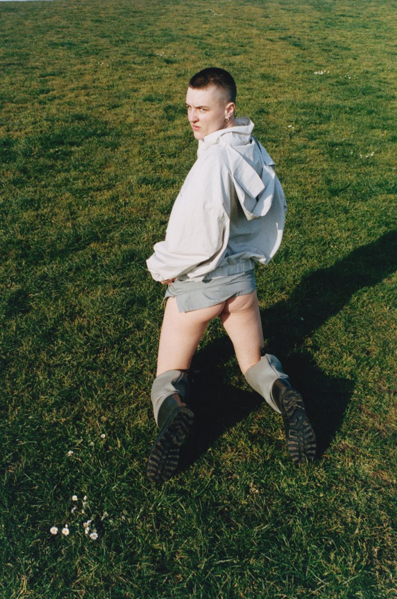 Adult poses kneeling on a grass lawn with a hoodie and boots, looking over their shoulder at the camera during the daytime.