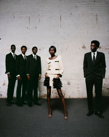 Fashion shot featuring an African-American woman in a skirt suit, posed with a group of men in black suits against a brick wall backdrop.
