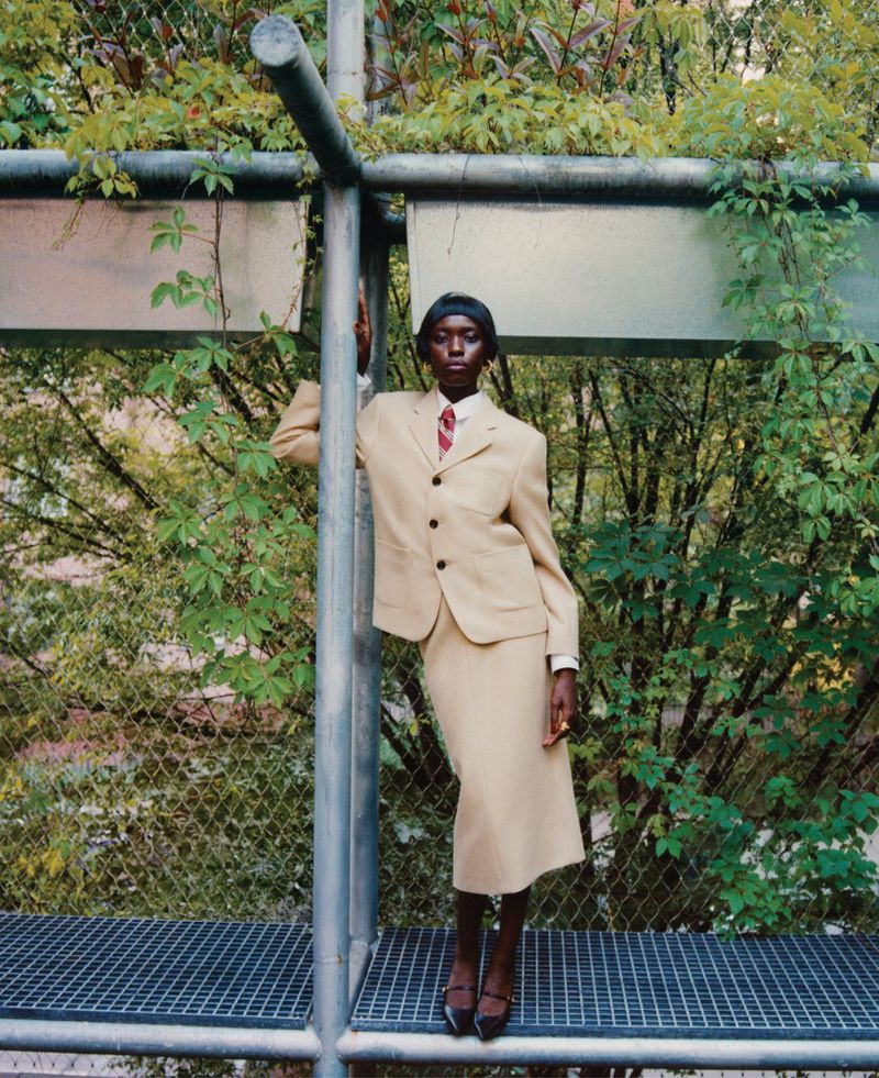 Elegant woman in a tan skirt suit poses outdoors near green foliage and a metal structure, highlighted by natural light and a fashion-forward aesthetic.