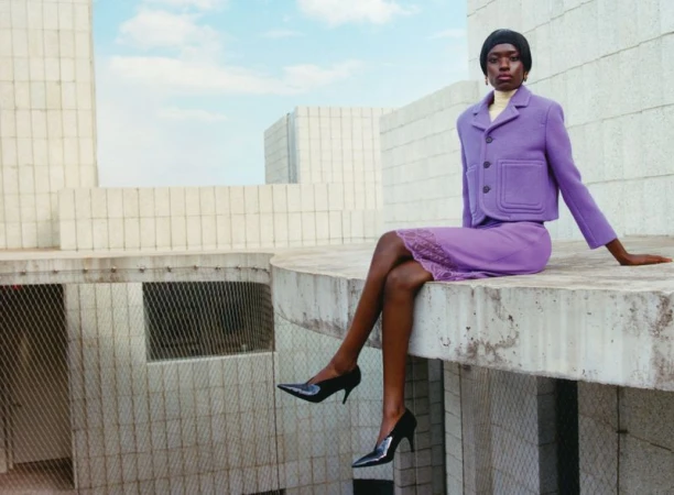 A black female fashion model poses sitting on a concrete structure, wearing a purple skirt suit with black heels, buildings in the background.