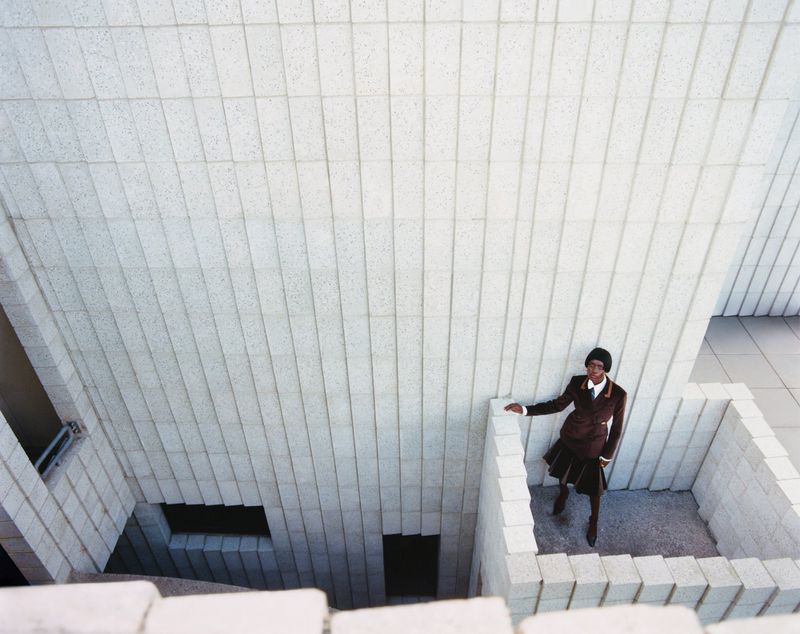 High-angle shot of an adult woman in a brown suit standing in a concrete architectural space with geometric walls and steps.