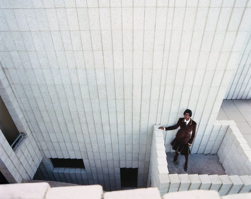 High-angle shot of an adult woman in a brown suit standing in a concrete architectural space with geometric walls and steps.