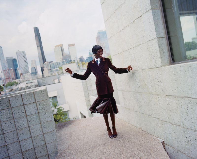 A fashionable African woman in a brown suit smiles on a rooftop with a cityscape backdrop, exuding confidence and elegance.