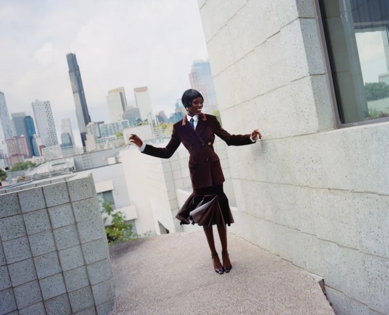 A fashionable African woman in a brown suit smiles on a rooftop with a cityscape backdrop, exuding confidence and elegance.