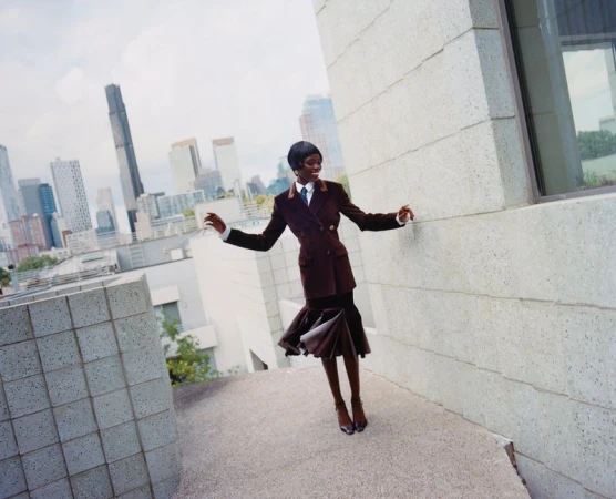 A fashionable African woman in a brown suit smiles on a rooftop with a cityscape backdrop, exuding confidence and elegance.
