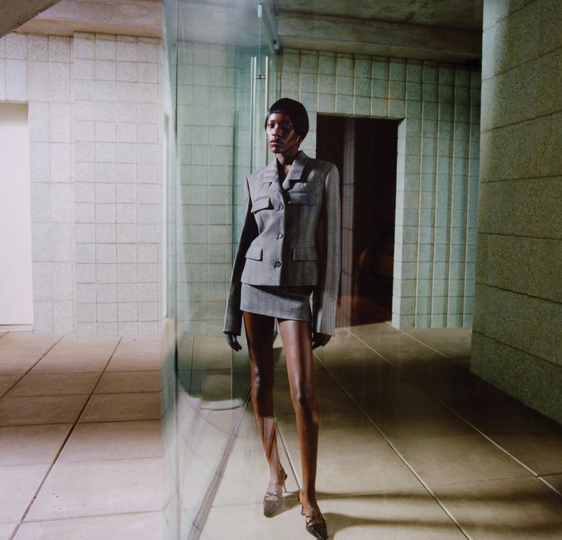 Woman in a grey skirt suit poses indoors with neutral tile walls and floor. She is centered and facing forward in a fashion shot.