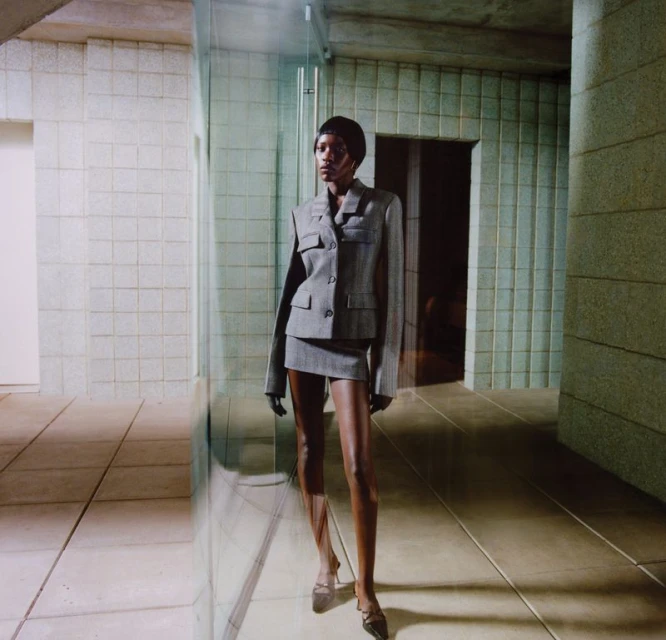 Woman in a grey skirt suit poses indoors with neutral tile walls and floor. She is centered and facing forward in a fashion shot.