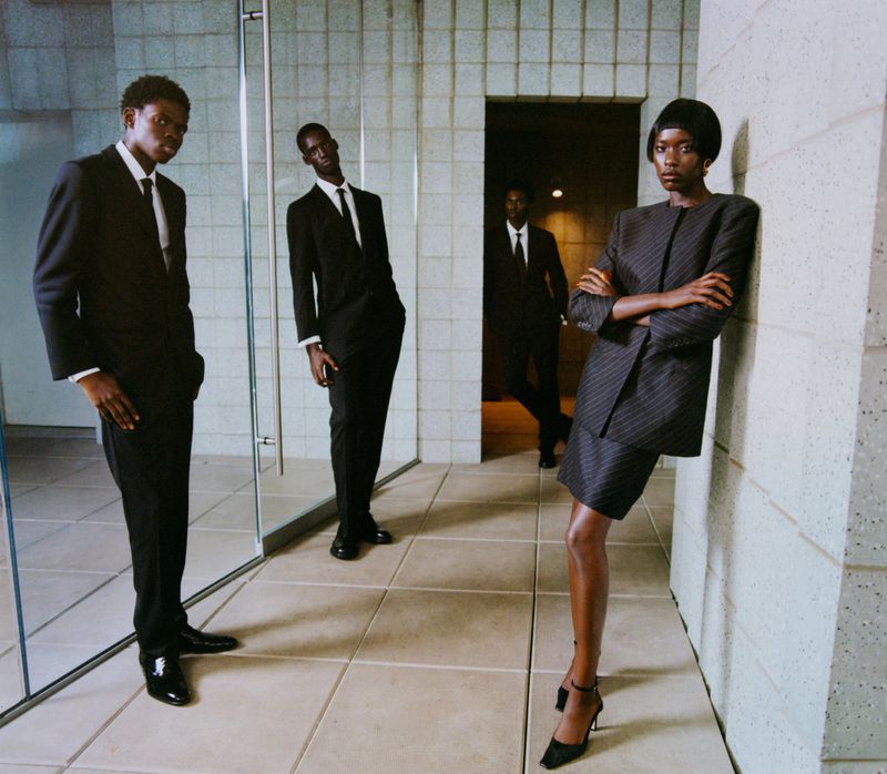 Three African American men in dark suits stand on a tiled floor, with a woman in a striped suit leaning against a wall.