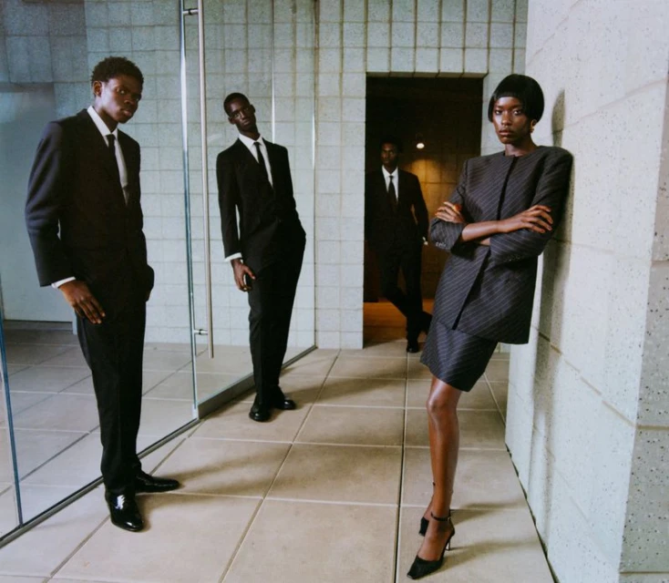 Three African American men in dark suits stand on a tiled floor, with a woman in a striped suit leaning against a wall.