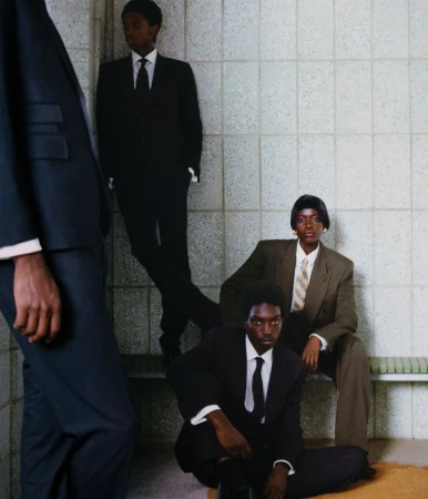 Black models wearing tailored suits and ties pose against a tiled wall in a corporate-inspired editorial setting.