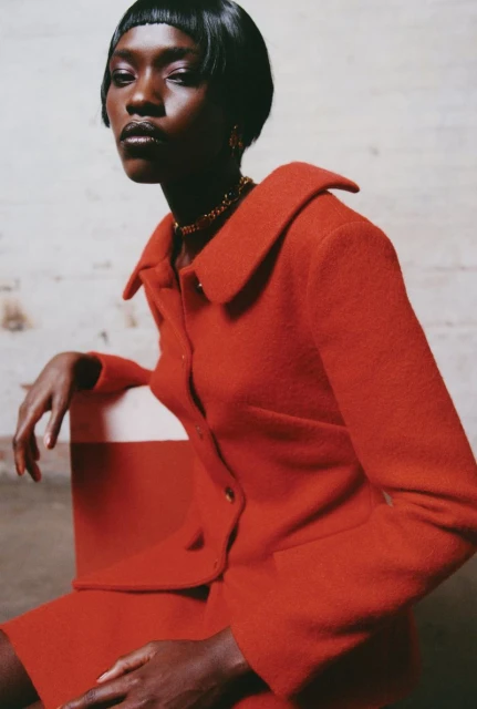 Fashion shot of a dark-skinned woman in an orange blazer and skirt, posing in front of a white wall. Professional studio lighting.