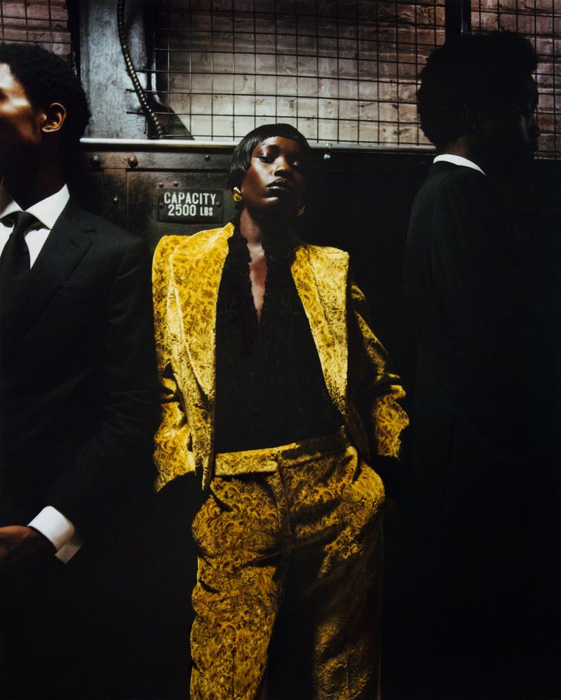 Fashion shot of an African American woman in a gold patterned suit, posing with two men in suits in a dark industrial setting.