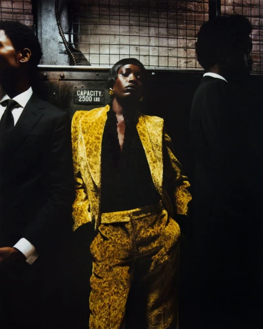 Fashion shot of an African American woman in a gold patterned suit, posing with two men in suits in a dark industrial setting.