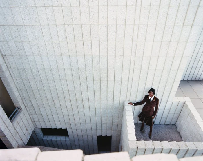 Model standing on a tiled balcony against a textured stone building wall, shot from a high angle.