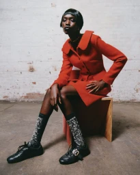 Model in a red coat and black shoes seated on a wooden block against a textured brick wall