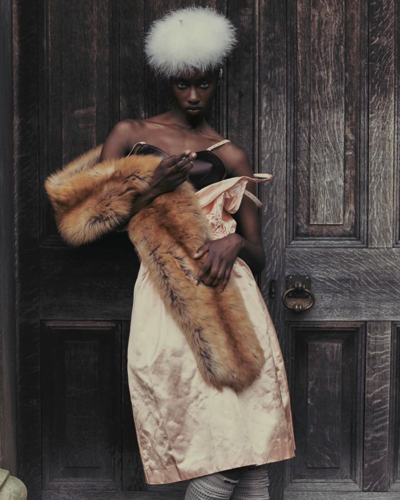 Fashionable black woman poses in peach dress and fur stole against weathered wooden door with white fuzzy hat, a study in texture and style.