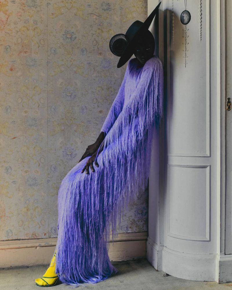 Adult female fashion model in a long-sleeve, fringed periwinkle gown and yellow heels, posing against a patterned wall next to a white corner cabinet.