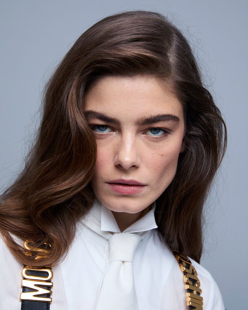 Portrait of adult woman with long brown hair, blue eyes, and wearing a white shirt with Moschino suspenders against a light gray backdrop.