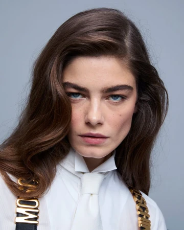 Portrait of adult woman with long brown hair, blue eyes, and wearing a white shirt with Moschino suspenders against a light gray backdrop.